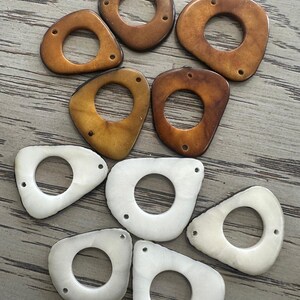 May include: A collection of irregularly shaped beads in shades of brown and white. Each bead has a central hole and two smaller holes for stringing. The beads vary in color from light tan to dark brown and white.