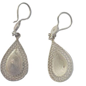 Puede incluir: Un par de pendientes plateados con forma de lágrima. Los pendientes presentan un delicado diseño de filigrana, con un centro en espiral y un borde estampado. Los ganchos son sencillos y curvados. Los pendientes se muestran sobre un fondo blanco.