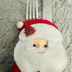 May include: A red and white Santa Claus-themed cutlery holder with a green Christmas tree design. The holder is made of felt and has a white beard and a red hat. A silver fork and knife are placed in the holder.