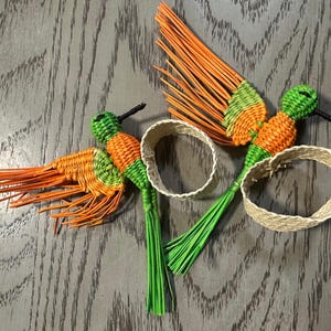 May include: Two handcrafted hummingbird ornaments. The birds are woven with orange, green, and black materials. Each bird has a long, orange tail and a green body. Two woven beige rings are also in the image.