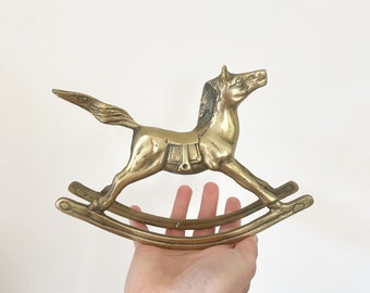 Puede incluir: Una figurita de caballo de balancín de latón de color dorado. El caballo está en posición de galope con la cabeza girada hacia un lado. La base de balancín está hecha de dos piezas curvas de latón.