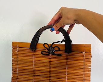 May include: A brown bamboo handbag with a dark brown wooden handle and a black bow with a decorative button in the centre.
