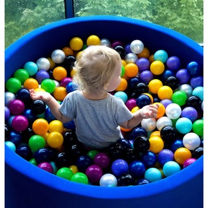 May include: A young child sits in a blue ball pit filled with colorful plastic balls. The child is wearing a grey shirt and has blonde hair. The ball pit is surrounded by a blue rim. The text '@flyingbubbles.lt' is visible in the bottom right corner of the image.