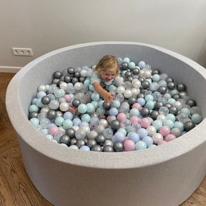 May include: A child plays in a gray ball pit filled with pastel blue, pink, and silver balls.