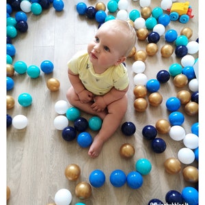 May include: A baby sits in a ball pit filled with blue, white, gold, and turquoise balls. The baby is wearing a yellow shirt with a pattern and is looking up.