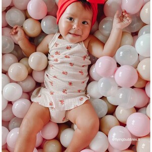 May include: A baby wearing a red bow headband and a white floral dress is playing in a ball pit filled with pink, white, and beige balls. The text '@flyingbubbles.lt' is visible in the bottom right corner of the image.