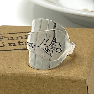 May include: A silver ring made from a spoon, with a floral pattern. The ring sits on a brown cardboard box with the text "Funk Vint". A white teacup and saucer are in the background.