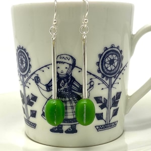 May include: A pair of silver earrings with green coffee bean charms hanging from a white teacup with a blue and white floral design.