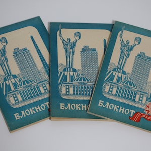 Può includere: Tre quaderni di carta blu con un design vintage che presenta un monumento, un edificio e una stella rossa. I quaderni hanno la parola "БЛОКНОТ" stampata sul davanti.