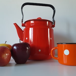 Puede incluir: Una tetera de esmalte rojo con lunares blancos y un asa negra, junto a una taza de esmalte naranja a juego con lunares blancos, y tres manzanas: una roja, una amarilla y una roja y amarilla.