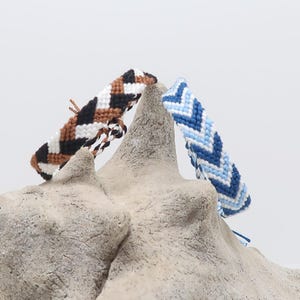 Peut inclure: Trois bracelets d'amitié faits à la main avec des motifs en chevrons marron, blanc et noir, et des nuances de bleu. Les bracelets sont exposés sur une sculpture texturée, de couleur claire, de forme organique sur fond blanc.