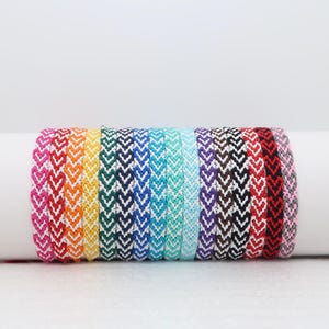 Peut inclure: Une collection de bracelets d'amitié colorés, chacun orné d'un motif en chevrons. Les bracelets présentent une variété de couleurs, notamment rose, orange, jaune, vert, bleu et violet. Ils sont présentés sur un fond blanc.