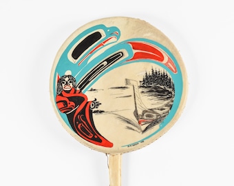 May include: Circular hand drum with a painted indigenous design. The artwork depicts a stylized eagle, a figure in red and black robes, and a boat on the water. The drum has a light-coloured wooden handle.