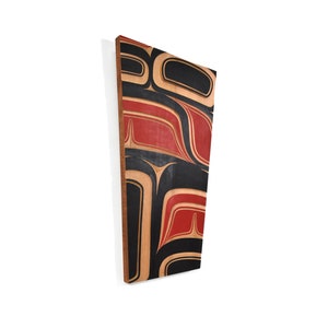 May include: A wooden wall art piece with a geometric design in black, red, and natural wood tones. The design is reminiscent of Northwest Coast Indigenous art.