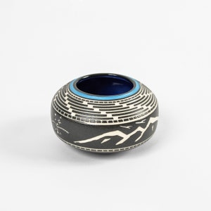 Peut inclure: Un petit bol en céramique noire avec un motif géométrique blanc et un bord bleu. Le bol présente une scène de montagne avec des oiseaux blancs volant au-dessus des montagnes.