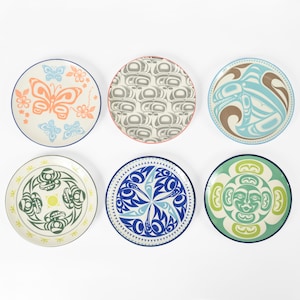May include: Six ceramic plates with different designs. The plates feature a variety of colors, including blue, green, yellow, orange, and white. The designs are inspired by Native American art and feature geometric patterns and stylized animals.