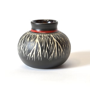 May include: A small, black ceramic vase with a white tree design and a red rim.
