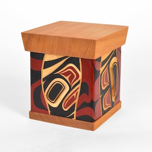 May include: A wooden box with a hinged lid. The box is decorated with a colorful Northwest Coast design featuring red, black, yellow, and white colors.