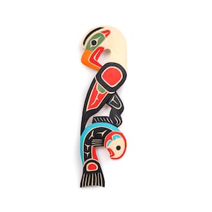 Puede incluir: Una talla de madera de un pájaro con un pez en el pico. El pájaro está pintado en negro, rojo, verde y blanco. El pez está pintado en negro, azul y rojo.