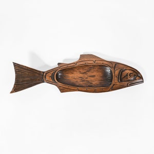 Puede incluir: Un plato de servir de madera con forma de pez con intrincados tallados. El plato es marrón con detalles negros y tiene un centro hueco para servir.