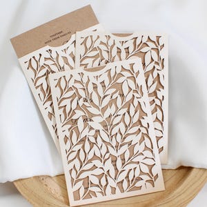 Peut inclure: Invitations crème découpées au laser avec un motif de feuilles, superposées sur une carte marron. La carte supérieure indique "TOGETHER WITH THEIR FAMILIES". Les invitations sont disposées sur un plateau en bois.