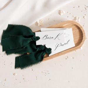 Può includere: Un set di segnaposto in carta bianca rettangolare con testo in calligrafia nera che riporta Bara K. e Pavel, ciascuno legato con un nastro di seta verde bosco scuro dai bordi sfrangiati, poggiato in un vassoio di legno chiaro ovale.