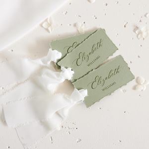 Op de afbeelding: Twee rechthoekige saliegroene papieren labels met gescheurde randen, elk voorzien van de naam Elizabeth Williams gedrukt in donkergroene inkt. Elk label is bevestigd aan een lang, wit zijden lint met gerafelde randen voor een gelaagd effect.