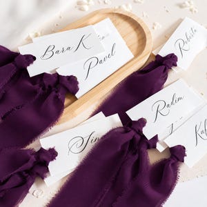 May include: A collection of white rectangular paper place cards featuring black calligraphy names including Bara K, Pavel, Roberta, Radim, and Sina. Each card is tied with a deep purple frayed chiffon ribbon, arranged on a light wood tray.