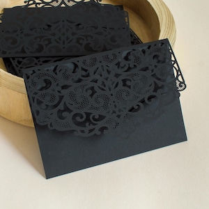 Könnte beinhalten: Schwarze Laser-geschnittene Umschläge mit einem zarten floralen Design. Die Umschläge sind in einer Holzschale gestapelt.