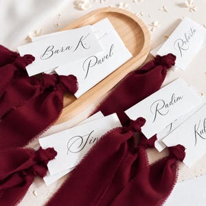 May include: A collection of white rectangular place cards featuring black calligraphy names including Bara K., Pavel, Roberta, Radim, and others. Each card is tied with a deep burgundy frayed-edge chiffon ribbon and rests on a wooden tray.