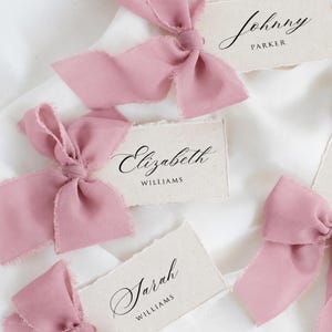 May include: Rectangular off-white paper place cards with deckled edges, each featuring a dusty rose chiffon ribbon tied in a bow. The cards are printed with black text reading Johnny Parker, Elizabeth Williams, and Sarah Williams in script.