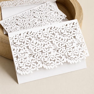 Op de afbeelding: Een witte papieren kaart met een ingewikkeld lasergesneden bloemenpatroon op de bovenste flap. Het ontwerp bestaat uit talloze kleine bloemen en bladeren die uit het karton zijn gesneden, wat zorgt voor een delicate, kantachtige uitstraling.