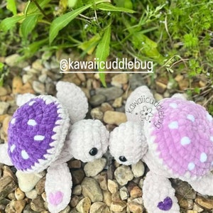May include: Two crocheted turtle plushies, one purple and one pink, with white polka dots on their shells. They are sitting on a bed of small rocks and green plants. The text "@kawaiicuddlebug" is visible in the image.