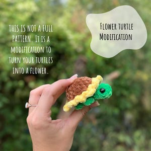 May include: A crocheted turtle with a brown shell, yellow flower petals, and green body. The turtle is being held in a hand. The text "FLOWER TURTLE MODIFICATION" is in a white speech bubble above the turtle. The text "THIS IS NOT A FULL PATTERN. IT IS A MODIFICATION TO TURN YOUR TURTLES INTO A FLOWER." is in the upper left corner of the image.