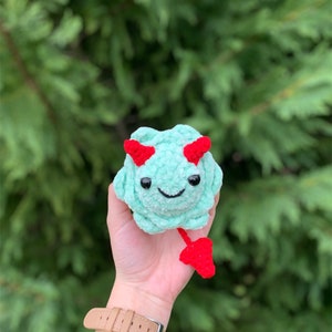 May include: A small crocheted plush toy in the shape of a green monster with red horns and a tail. The monster has a smiling face with black eyes and a red mouth.