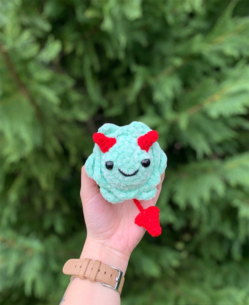 May include: A small crocheted plush toy in the shape of a green monster with red horns and a tail. The monster has a smiling face with black eyes and a red mouth.