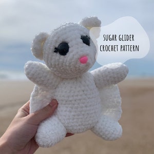 May include: White crocheted sugar glider plush toy with black eyes and a pink nose. The toy is being held in a hand with the beach in the background. The text "SUGAR GLIDER CROCHET PATTERN" is in a speech bubble above the toy.