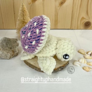 May include: A crocheted turtle toy with a purple and white shell. The turtle is sitting on a wooden slice with a white background. The text "@straightuphandmade" is visible below the turtle.