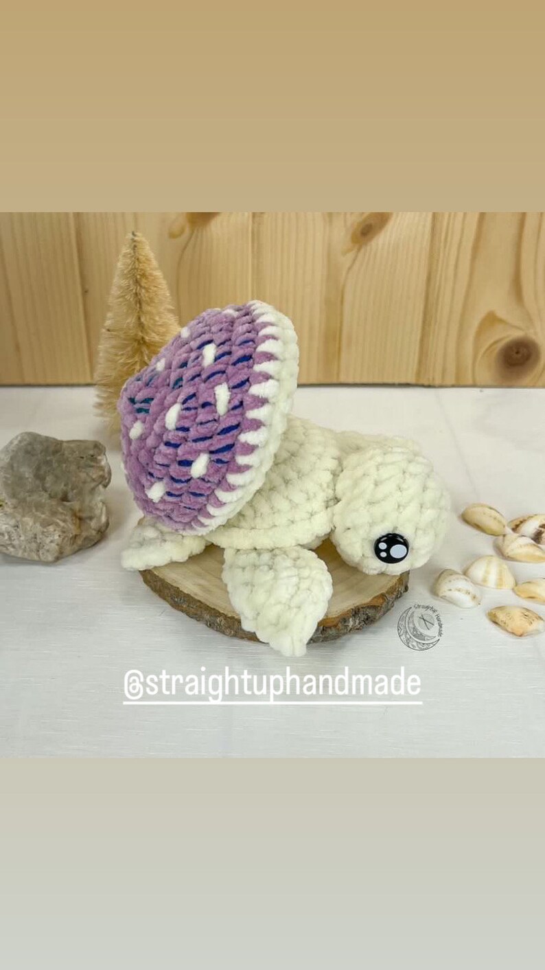 May include: A crocheted turtle toy with a purple and white shell. The turtle is sitting on a wooden slice with a white background. The text "@straightuphandmade" is visible below the turtle.
