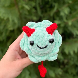 May include: A small, green crocheted plush toy with red horns and a tail. The toy has a smiling face with black eyes and a black stitched mouth.