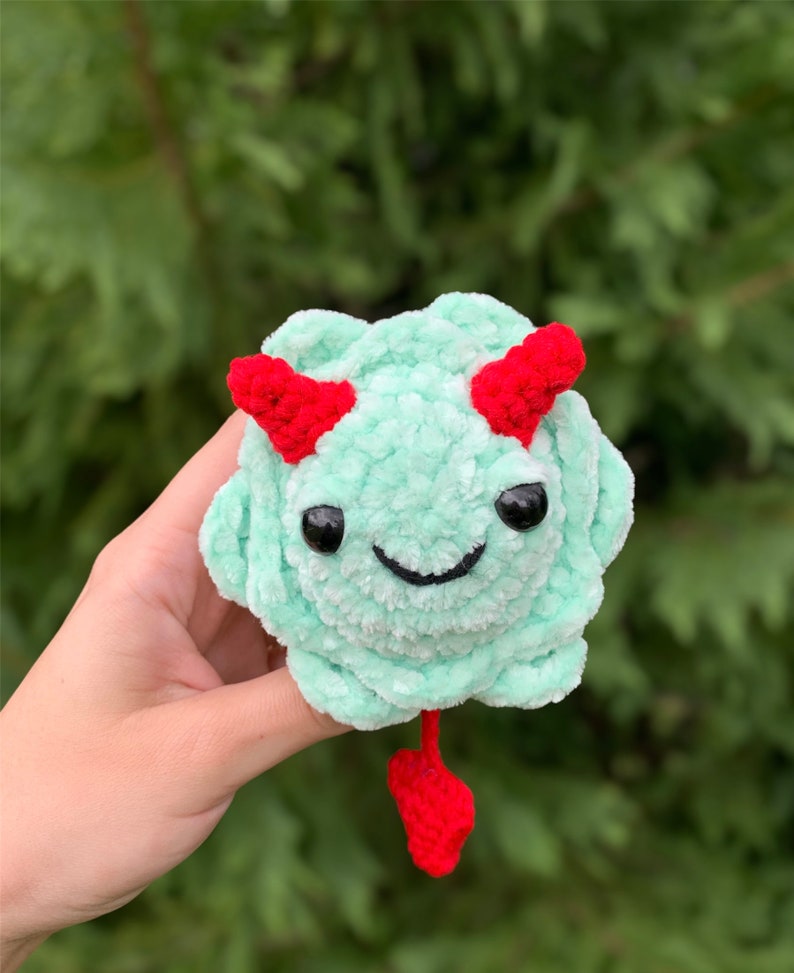 May include: A small, green crocheted plush toy with red horns and a tail. The toy has a smiling face with black eyes and a black stitched mouth.