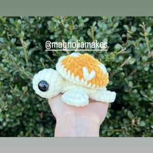 May include: A crocheted turtle with a white body and a brown shell with white accents. The turtle has large black eyes. The turtle is being held in a hand. The image includes the text "@magnoliamakes".
