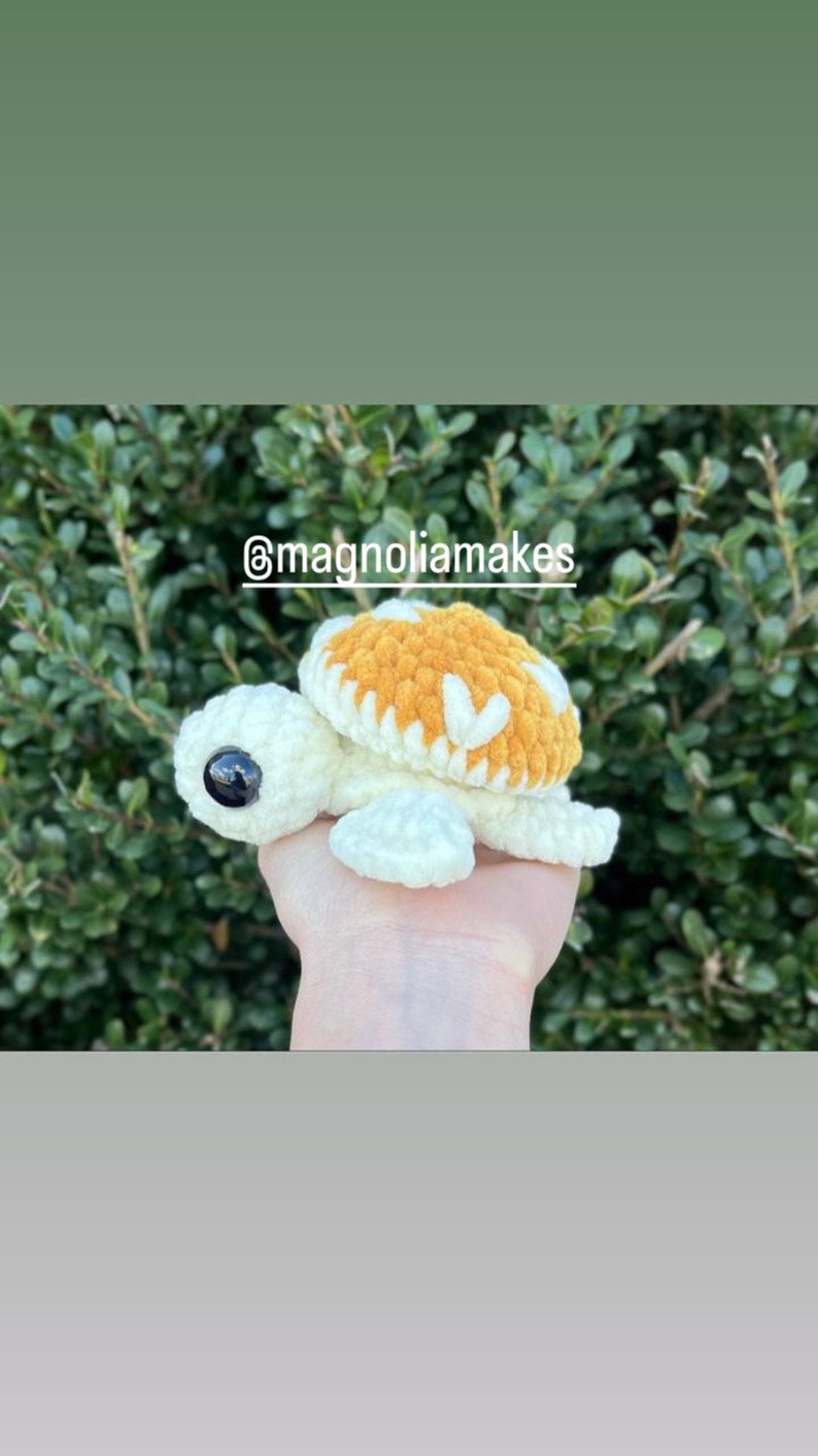 May include: A crocheted turtle with a white body and a brown shell with white accents. The turtle has large black eyes. The turtle is being held in a hand. The image includes the text "@magnoliamakes".