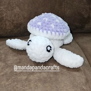May include: A crocheted stuffed turtle toy with a white body and a purple shell with white polka dots. The turtle has black eyes and a cute expression. The toy is sitting on a brown couch. The text "@mandapandacrafts" is visible in the image.