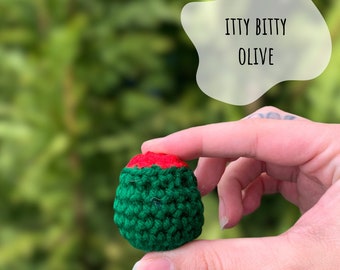 May include: A small crocheted olive, green with a red top, held in a hand. The text "ITTY BITTY OLIVE" is in a white speech bubble above the olive.