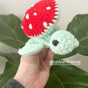 May include: A crocheted green turtle with a red and white mushroom shell. The turtle has black eyes and a small mouth. The turtle is being held in a hand.