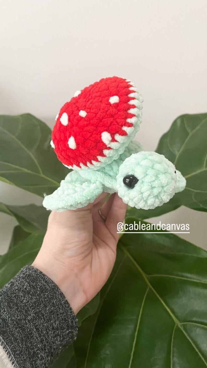 May include: A crocheted green turtle with a red and white mushroom shell. The turtle has black eyes and a small mouth. The turtle is being held in a hand.