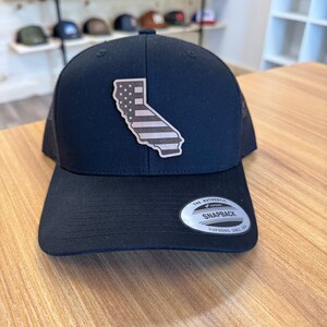 May include: Black snapback hat with a mesh back and a curved brim. The hat features a patch in the shape of California with a design of the American flag. The hat has a snapback closure and a circular tag that reads "The Authentic Snapback".