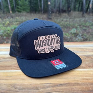 May include: A navy blue flat bill snapback hat with a mesh back. The hat features a patch with the words "FEEDING MOSQUITOS SINCE BIRTH" and a mosquito graphic. A small tag with the brand name is visible.