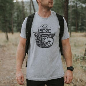 May include: A light gray t-shirt featuring a black graphic of a bear wearing a ranger hat. The text "I PUT OUT" is above the bear's head. The shirt is being worn in a forest setting.